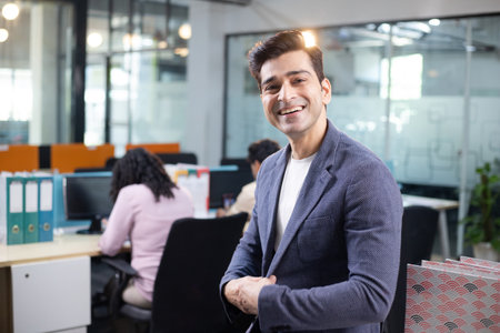 Portrait of smiling businessman standing with arms crossed in office with colleagues in backgroundの写真素材