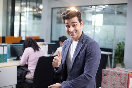 Portrait of smiling businessman giving thumbs up while colleagues working in background at officeの写真素材