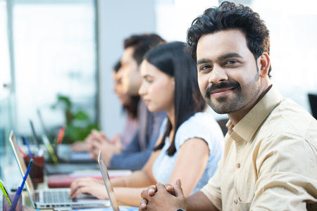 Portrait of smiling businessman working on laptop with colleagues in background at office