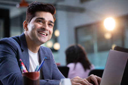 Portrait of smiling businessman using laptop at desk in creative office.