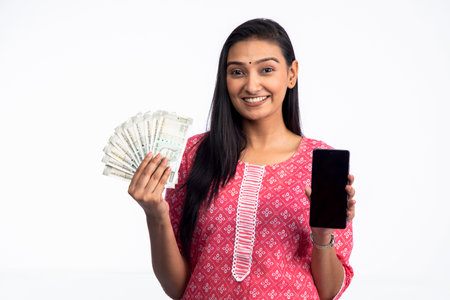 Indian woman showing indian currency notes while holding cellphone and looking at camera isolated...