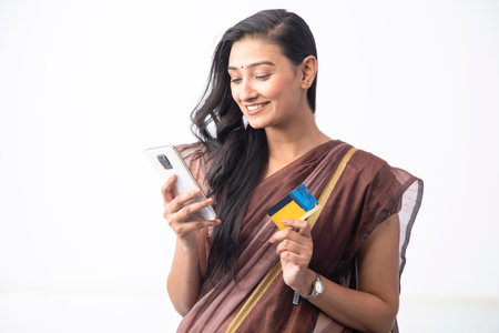Smiling Indian woman holding credit card and using mobile phone while standing against white...