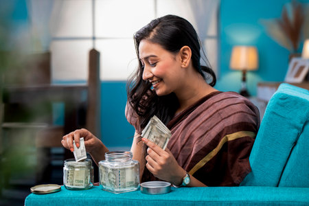 Young Indian woman counting money in a glass jar sitting on a sofa at home