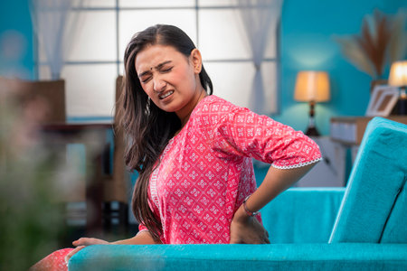 Asian woman suffering from backache while sitting on sofa at home.