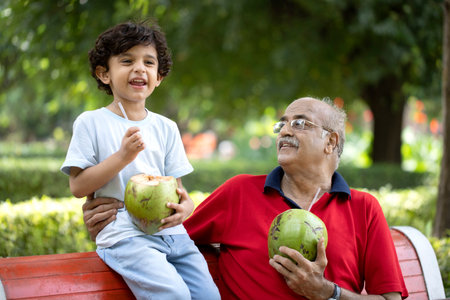 Grandfather and grandson sitting on bench in the park and eating coconut