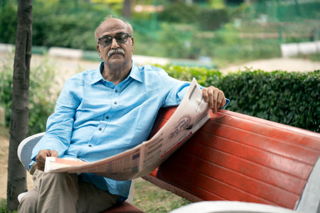 indian senior man reading newspaper while sitting on bench in public parkの写真素材