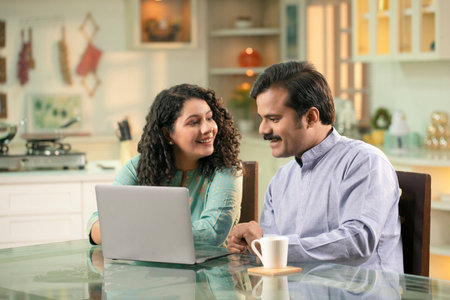 Smiling indian couple using laptop computer in kitchen at home.