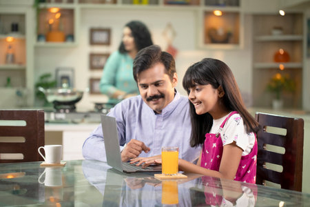 Indian father and daughter using laptop computer at table in kitchen at home