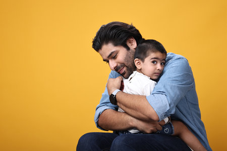 Indian father hugging his son while sitting on the floor over yellow backgroundの写真素材