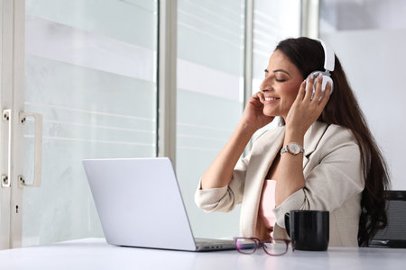 Beautiful young woman with laptop and headphones listening to music in officeの写真素材
