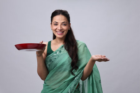 indian woman holding a plate and looking at the camera on grey backgroundの写真素材