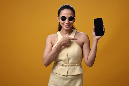 Portrait of a young beautiful woman showing mobile phone on yellow backgroundの写真素材