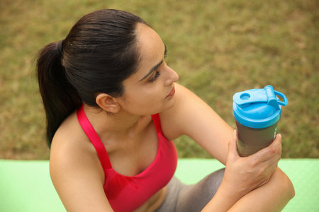 Young woman sitting on yoga mat and holding bottle of water, outdoors