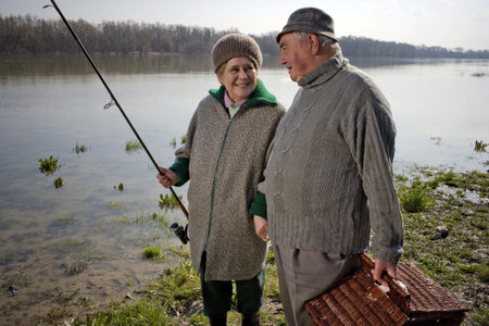 Senior couple carrying picnic basket and fishing rod by river, smiling