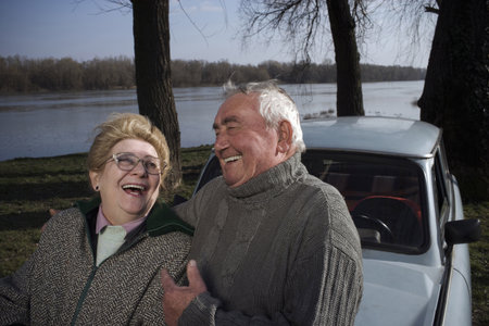 Senior couple standing by car on riverside, laughingの写真素材