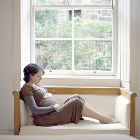Portrait Of Mid Adult Pregnant Woman Sitting On Window Seat Holding Stomachの写真素材