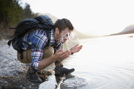 Young Man, Hiking, Crouching By Lake, Splashing Face With Waterの写真素材