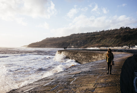 Rear View Of Young Woman Strolling Along 'The Cobb' Harbour Wall, Lyme Regis, Dorset, England