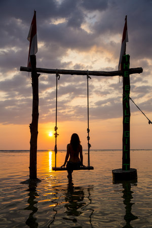 Silhouetted Female Tourist On Swing Watching Sunset Over Sea, Gili Trawangan, Lombok, Indonesiaの写真素材