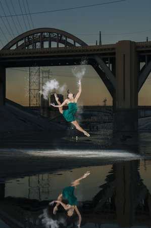 Female Ballet Dancer Leaping Whilst Releasing Powder Explosion Above River At Sunset, Los Angeles, Usaの写真素材