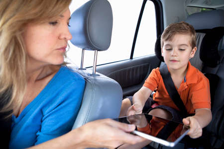 Mother passing digital tablet to son in back seat of car