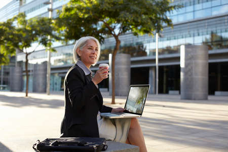 Businesswoman sitting outside using laptop and drinking hot drinkの写真素材