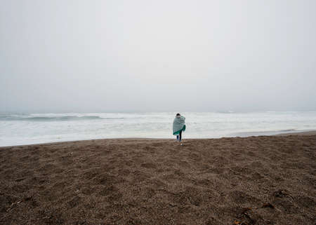Young woman wrapped in blanket walking on misty beachの写真素材