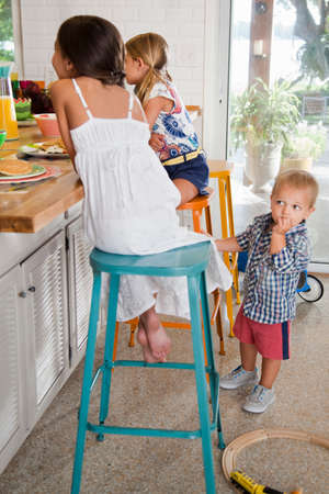 Two sisters at kitchen breakfast bar with toddler brother watchingの写真素材