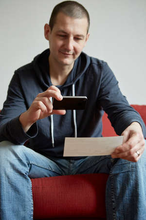 Mid adult man sitting on sofa photographing paper with smartphoneの写真素材