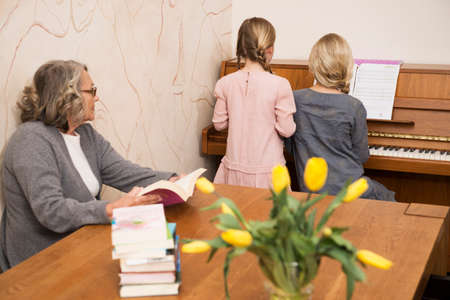 Two sisters playing piano watched by grandmotherの写真素材