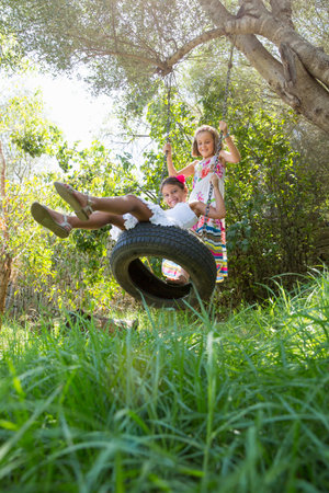 Two girls sitting and standing on tree swing in gardenの写真素材