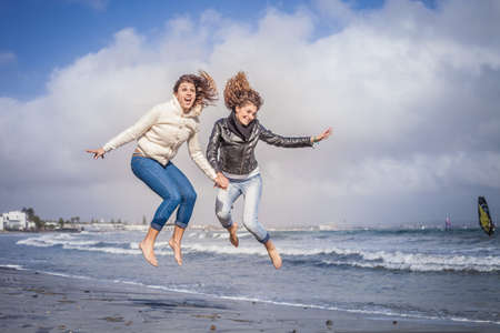 Two young women friends holding hands and jumping on beachの写真素材