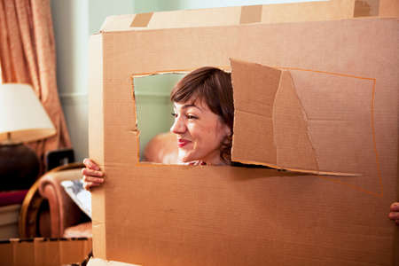 Mid adult woman peeking out of cardboard box window in living roomの写真素材