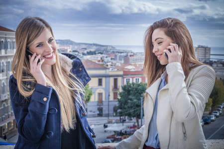 Two young women on rooftop talking on smartphones, Cagliari, Sardinia, Italy