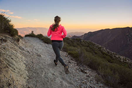 Rear view of young woman trail running up dirt track at dusk on Pacific Crest Trail, Pine Valley,...