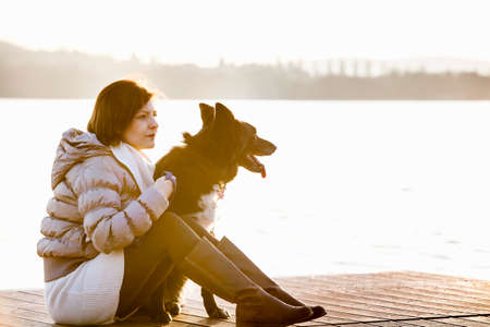 Mid adult woman and her dog sitting on sunlit lake pierの写真素材