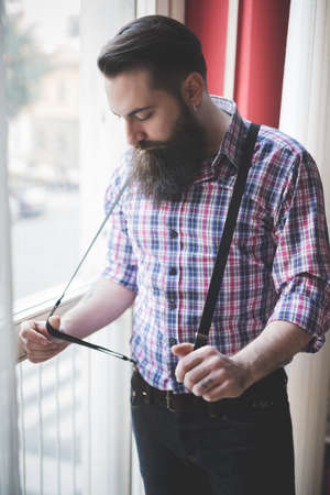 Young bearded man pulling braces by windowの写真素材