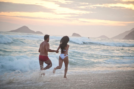 Mid adult couple running along beach as tide comes in, hand in hand, rear viewの写真素材
