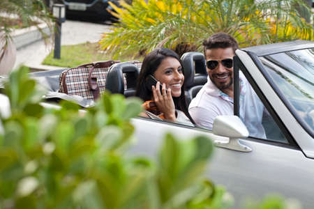 Mid adult couple in convertible car, woman using smartphone