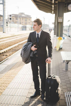 Young businessman commuter standing on railway platform with suitcase.の写真素材