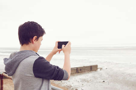Teenage boy photographing sea on smartphone, Southend on Sea, Essex, UKの写真素材