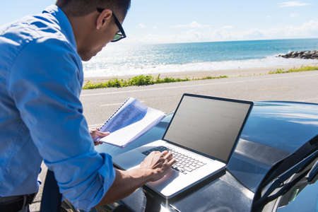 Young businessman at coast parking lot typing on laptop whilst reading paperwork