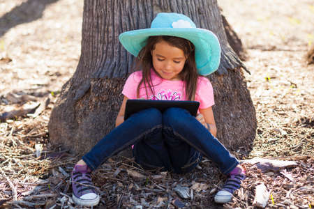 Cute girl leaning against tree trunk using digital tablet