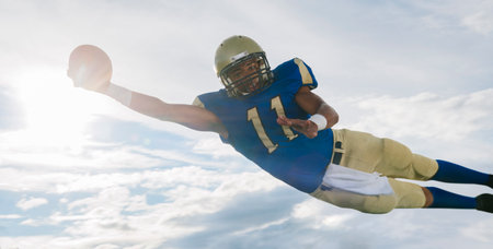 Male teenage American football player catching ball mid air against sunlit sky