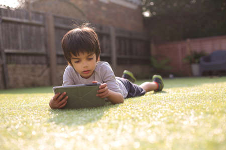 Boy in garden lying on front on grass looking down using digital tabletの写真素材
