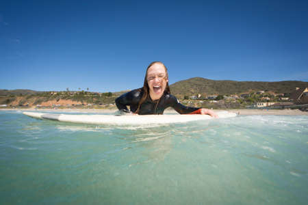 Front view of young woman lying on surfboard looking at camera sticking out tongue. Los Angeles, California, USAの写真素材