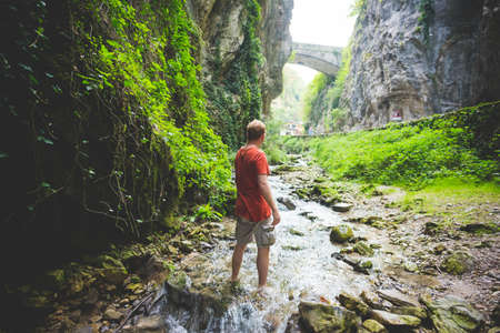 Man standing in stream, rock hills and stone bridge in background, Garda, Italy