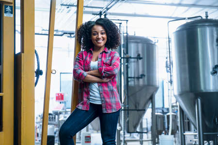 Mid adult woman in brewery leaning against yellow fork lift truck, arms folded looking at camera smilingの写真素材