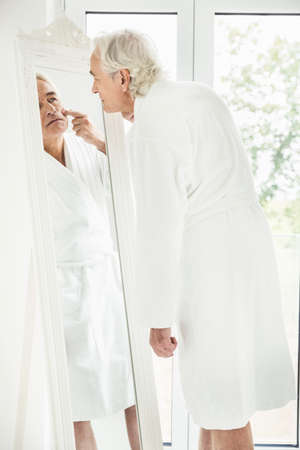 Grey haired senior man wearing bathrobe staring at himself in mirrorの写真素材