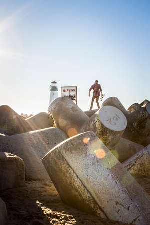 Young man holding skateboard, standing on breakwater near lighthouse, rear viewの写真素材